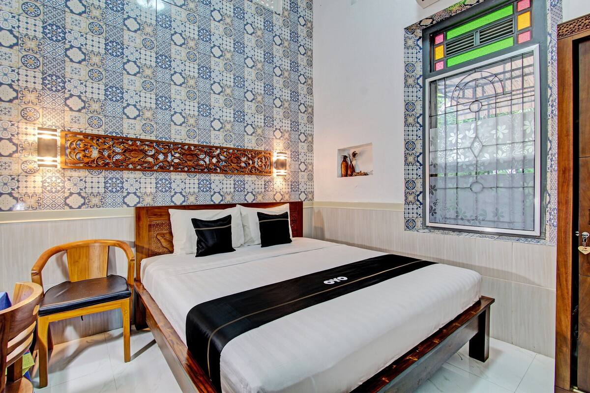 A neatly arranged bedroom with a bed, a wooden chair, a decorative wall panel, and a window with a view.