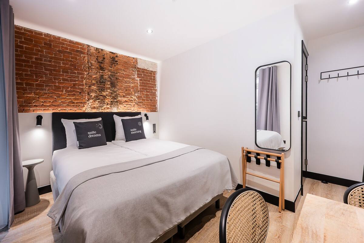 Habitación de hotel moderna con pared de ladrillo decorativa, una cama grande con dos almohadas, una mesita de noche, un espejo y una pequeña mesita auxiliar de madera.