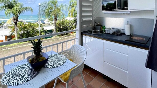 A modern kitchen with white cabinets, a black countertop, and a ceiling fan. A dining table with a pineapple and a bowl of fruit is set outside on a balcony with a view of the ocean and palm trees. - Chez Valerie ET Pascal (Tartane)