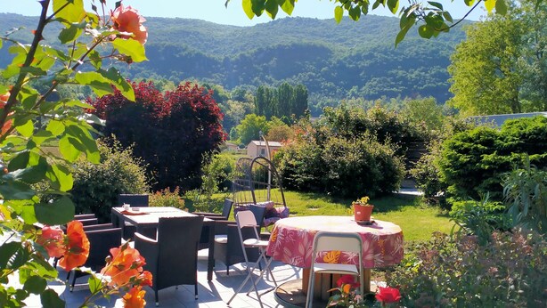 An outdoor patio with a table and chairs, surrounded by lush greenery and a view of a hilly landscape. - Chambre d'Hôtes l'Hôsting Noix (Hostun)
