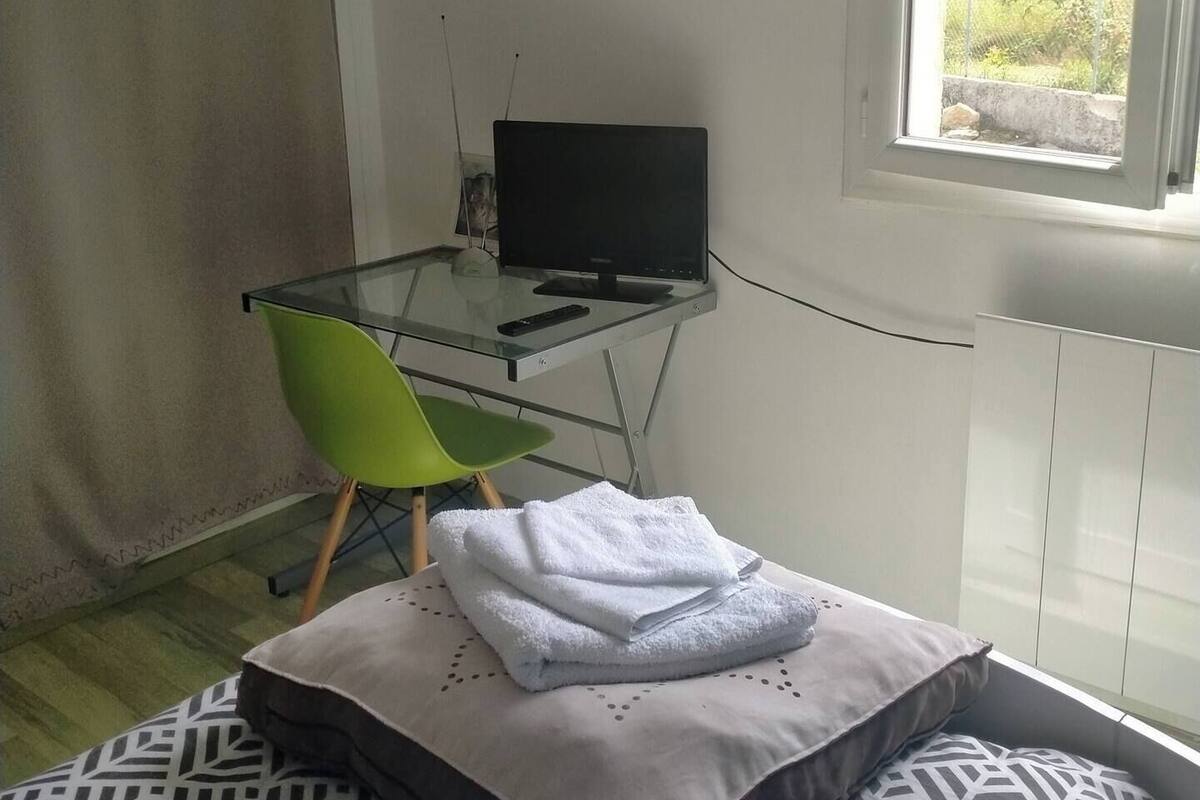 A bedroom with a bed, a desk with a computer, a green chair, and a window with a view of greenery.