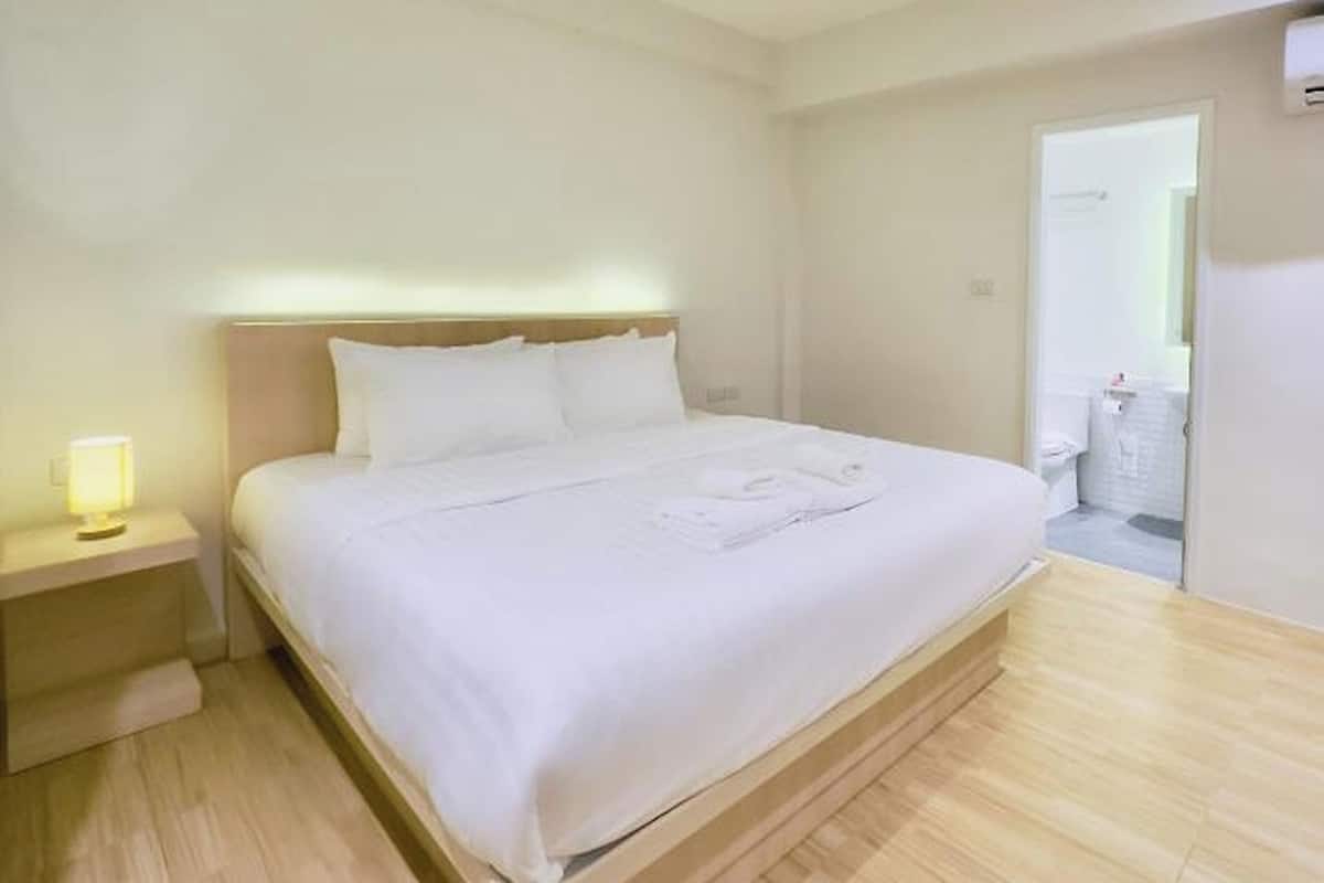 Cama bem arrumada com lençóis brancos em um quarto com piso de madeira e uma mesa de cabeceira com abajur.