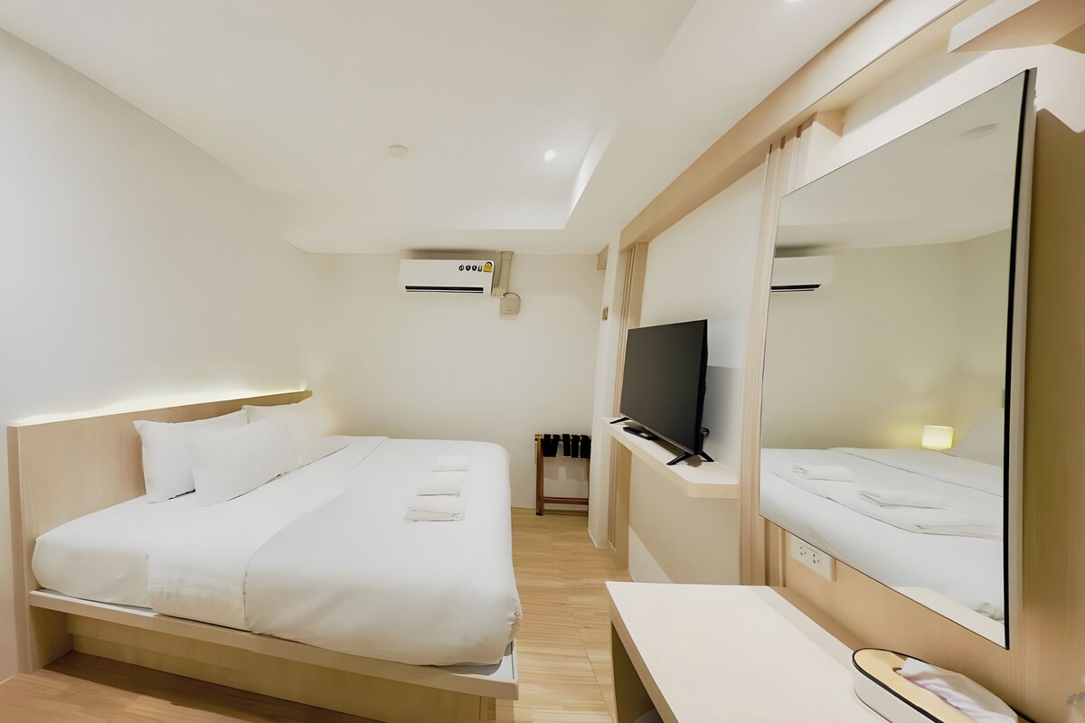 A hotel room with a bed, a TV, a mirror, and an air conditioning unit.