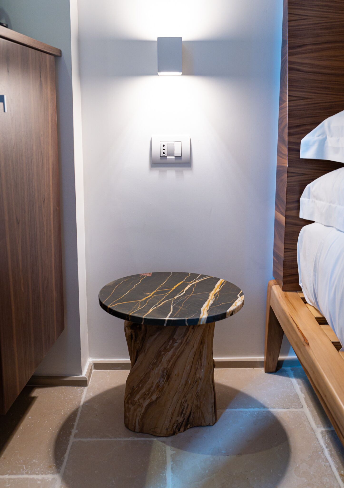 A modern bathroom with a marble vanity, a round table with a marble top, a wooden bench, and a wall-mounted light fixture.