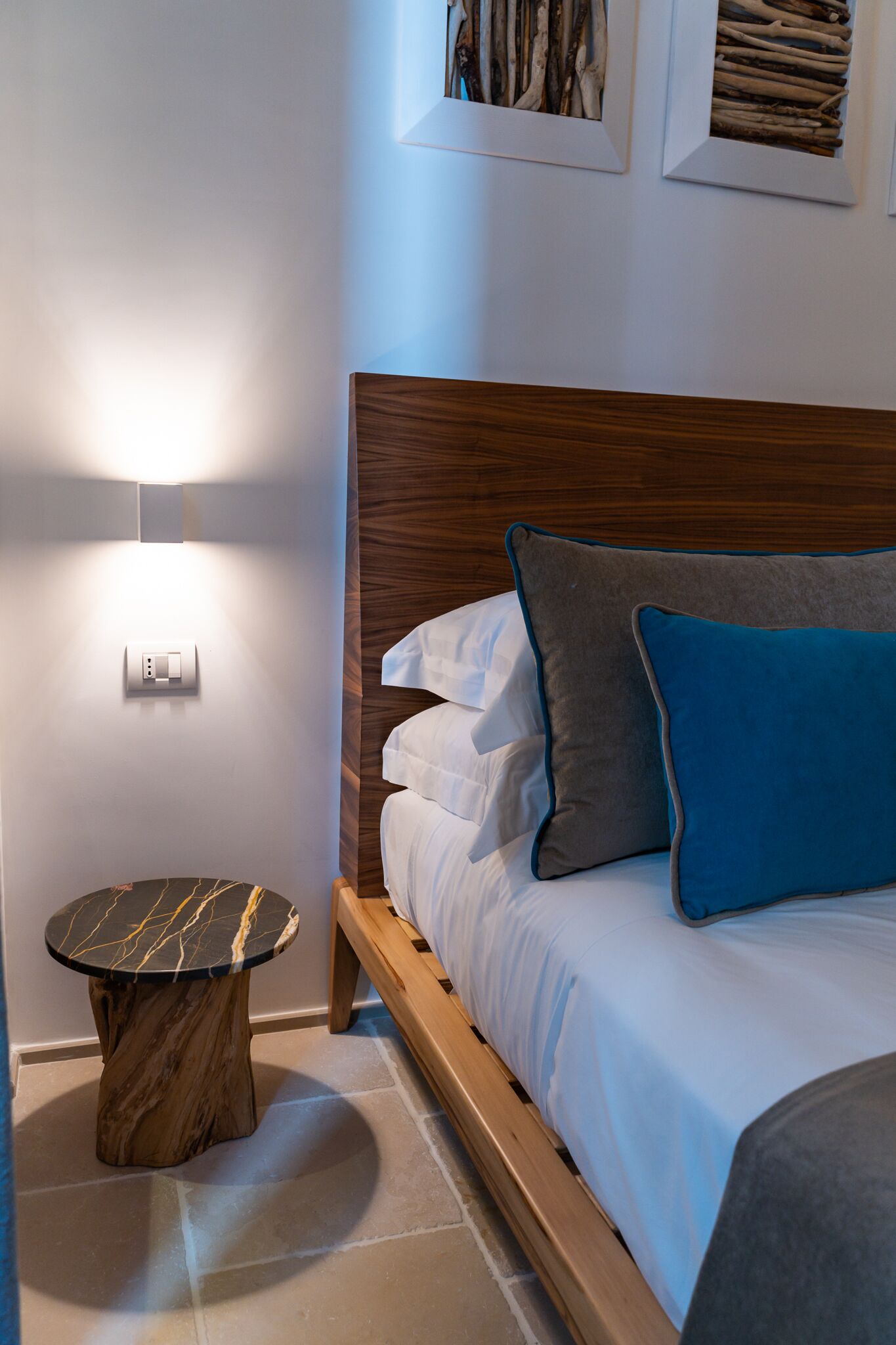 A neatly made bed with white and blue pillows, a round wooden side table, and a wall-mounted light fixture.