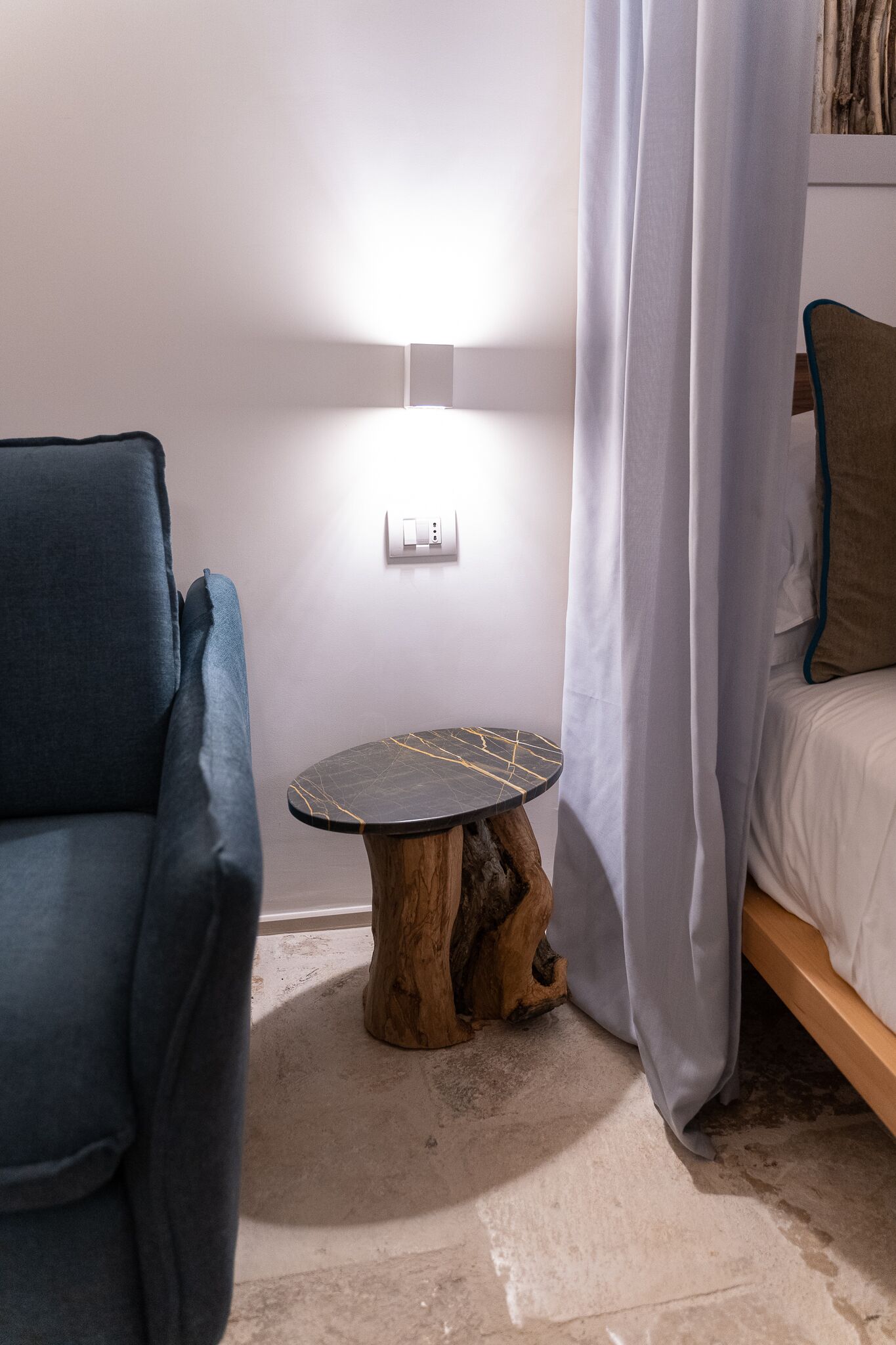 Een kamer met een blauwe fauteuil, een kleine ronde tafel met een marmeren blad, een houten bijzettafeltje, een bed met wit beddengoed en een wit gordijn.