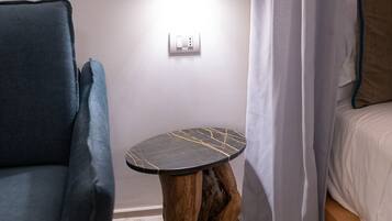 Een kamer met een blauwe fauteuil, een kleine ronde tafel met een marmeren blad, een houten bijzettafeltje, een bed met wit beddengoed en een wit gordijn.