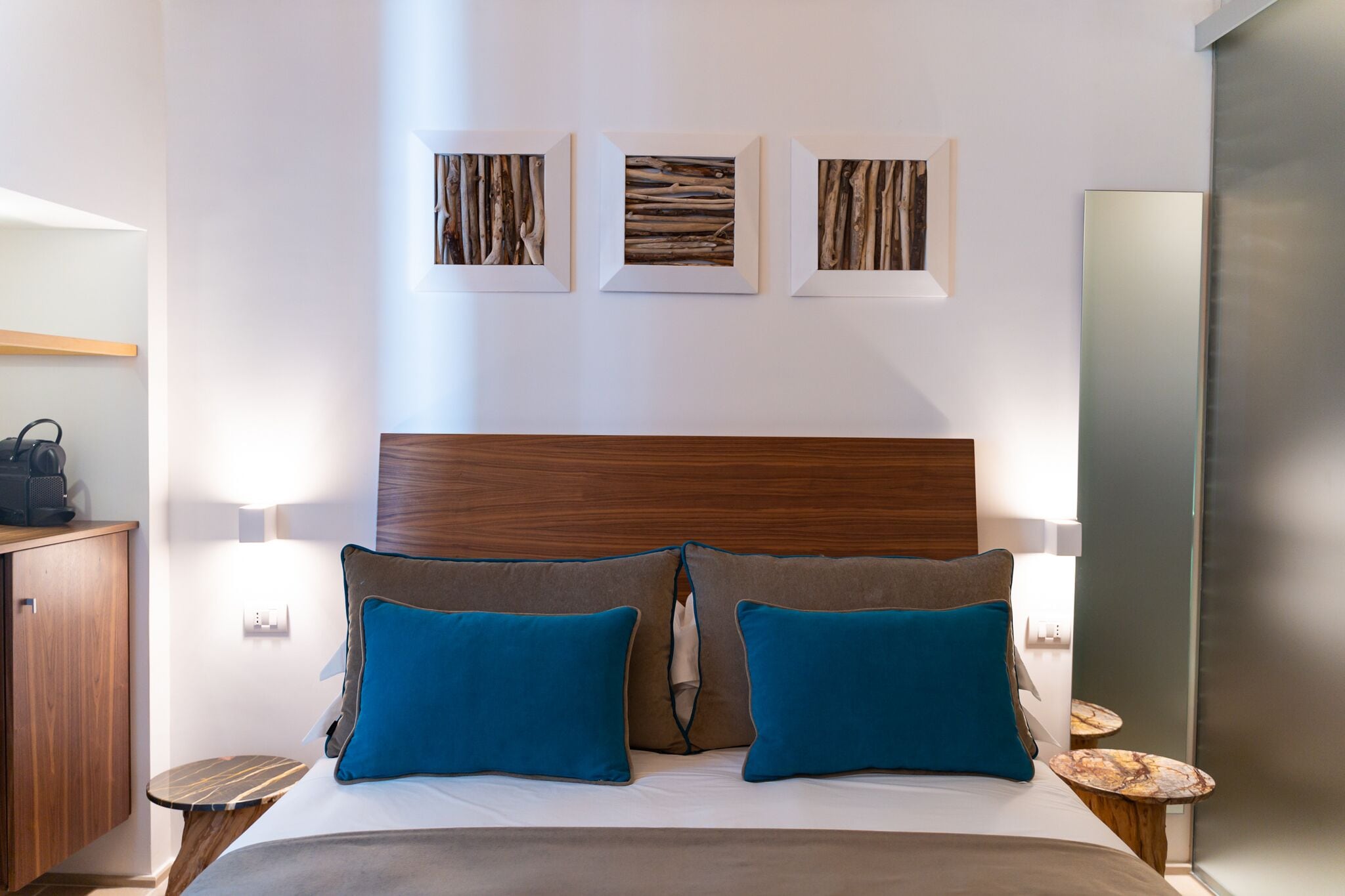 Ein modernes Hotelzimmer mit einem hölzernen Kopfteil, einem Bett mit blau-braunen Kissen, einem kleinen Nachttisch aus Holz und drei gerahmten Kunstwerken an der Wand.