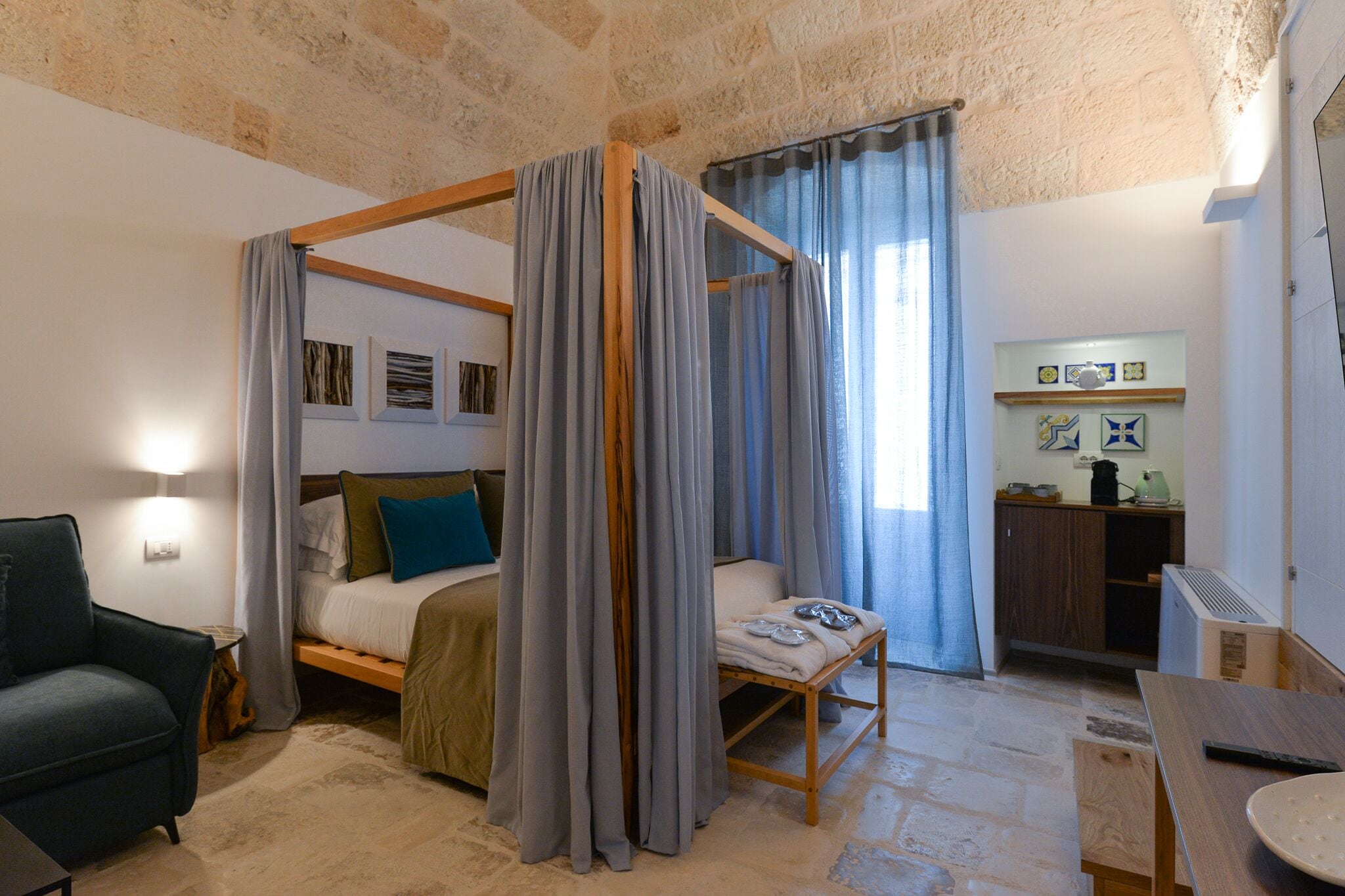 Una cama con dosel, cortinas grises, un banco de madera, una mesita y una silla en una habitación con paredes de piedra y una kitchenette.