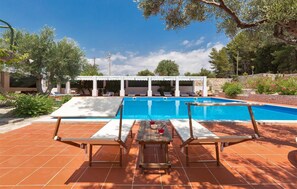 A swimming pool with lounge chairs and a table, surrounded by greenery and a stone wall. - 836 Masseria Colosso - Pajara (Patu)