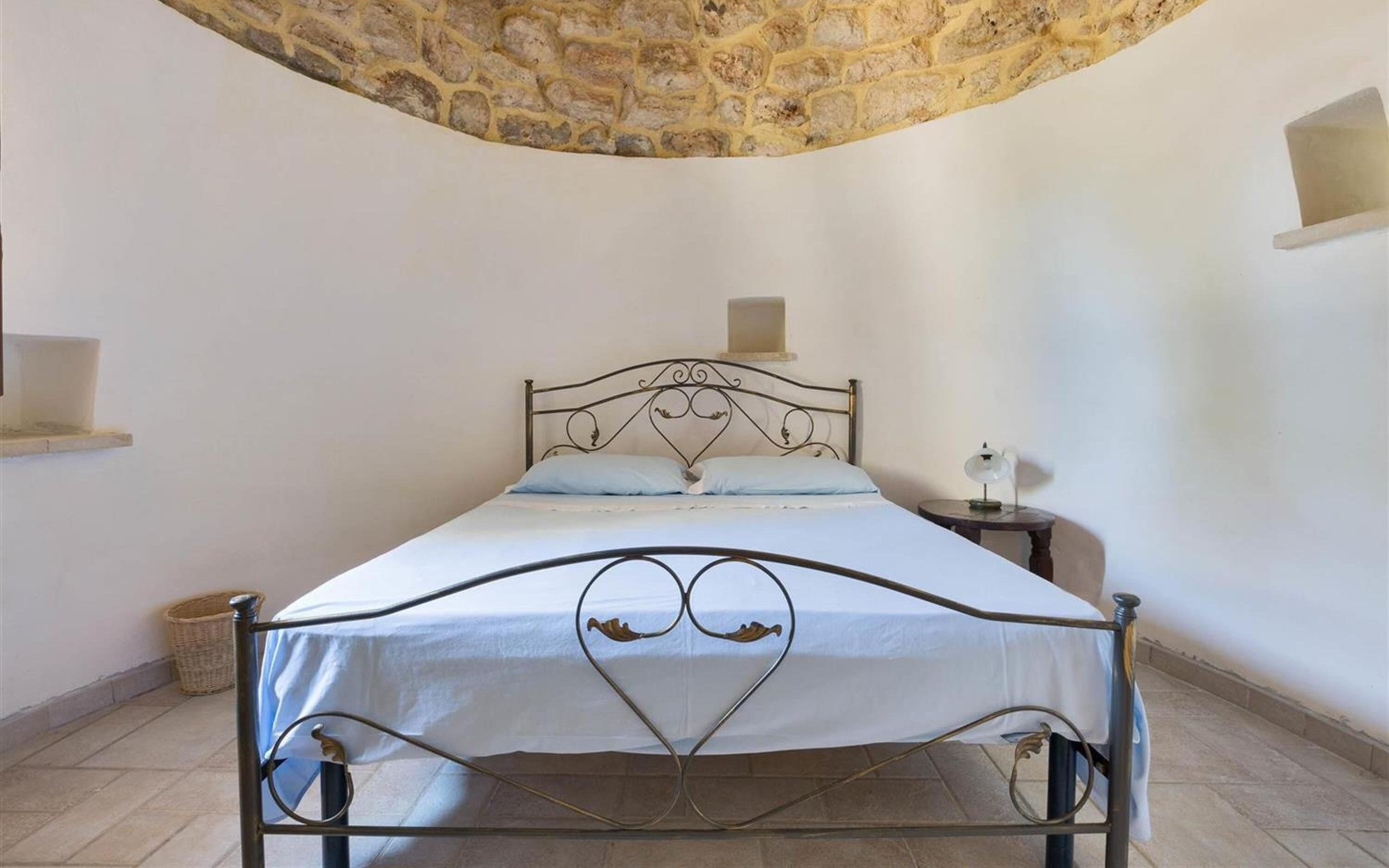 A bed with a heart-shaped metal frame, a stone wall, and a small bedside table with a lamp.