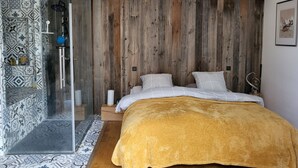 Een moderne slaapkamer met een douchecabine van glas, patroontegels, een houten hoofdbord en een mosterdgele deken.