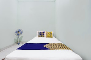 Une petite chambre avec un lit simple, recouverte d’une courtepointe blanche, agrémentée de deux coussins à motifs, d’une couverture bleue et jaune, et équipée d’un ventilateur sur pied.
