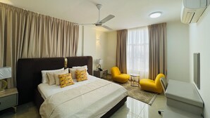 Een hotelkamer met een bed, een plafondventilator, een klein tafeltje, twee gele stoelen en een raam met gordijnen.