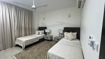 A hotel room with two beds, a ceiling fan, a large window with curtains, and a wall-mounted air conditioner.