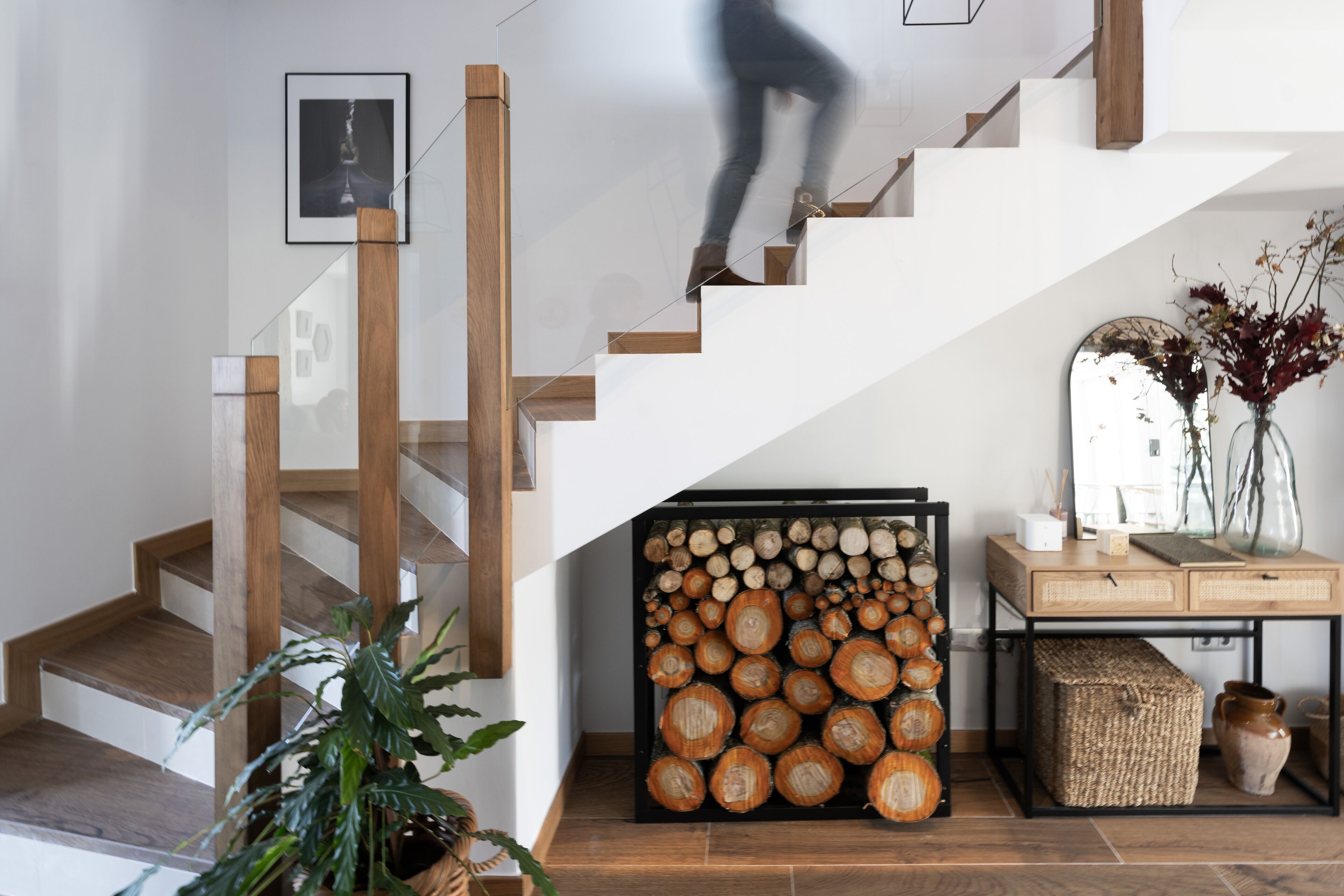Una escalera con escalones de madera y barandillas de vidrio, un perchero de troncos en la pared, y una pequeña mesa con un florero y adornos.