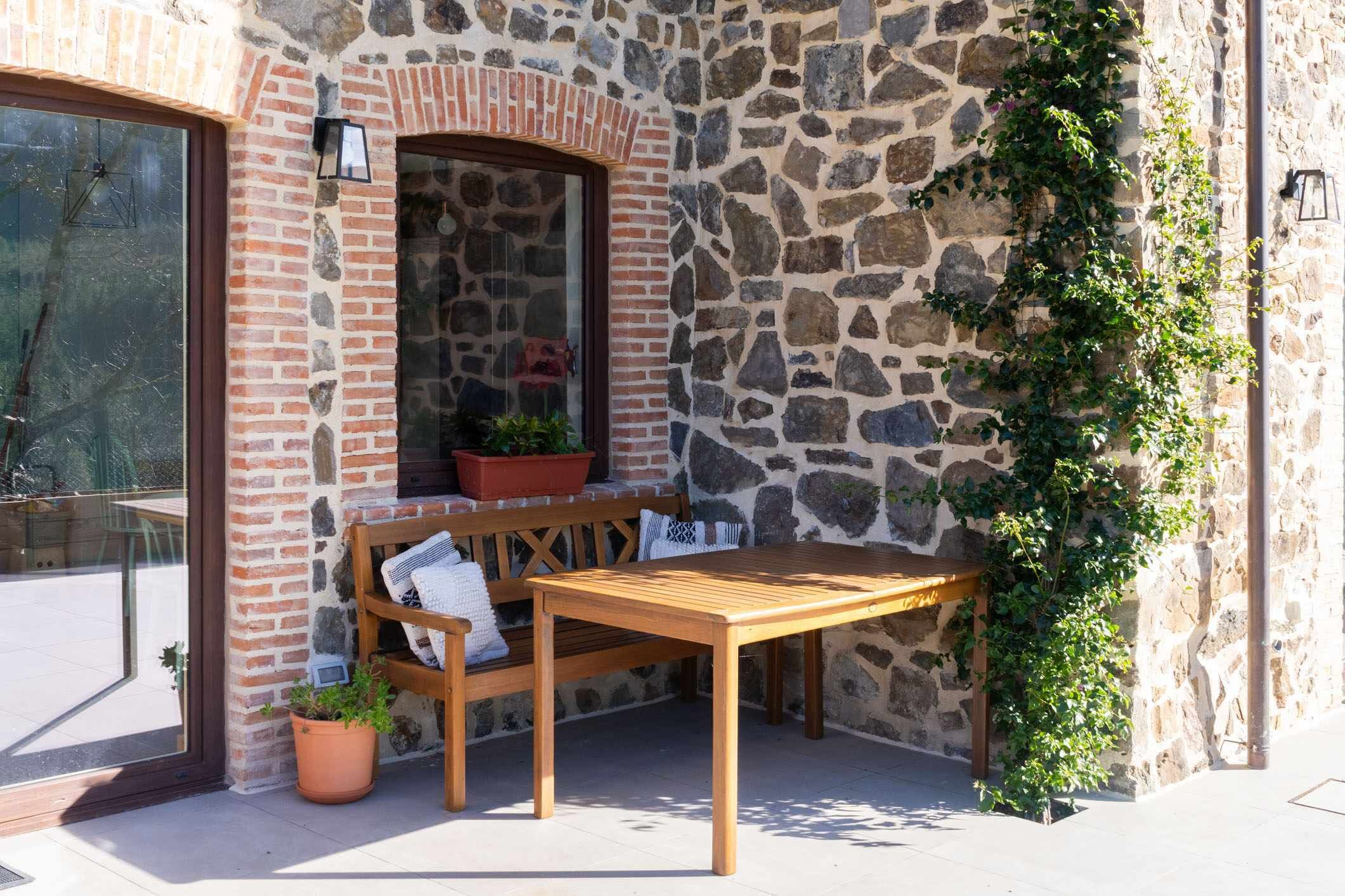 Un muro de piedra con un banco y una mesa de madera, una planta en maceta y una ventana.