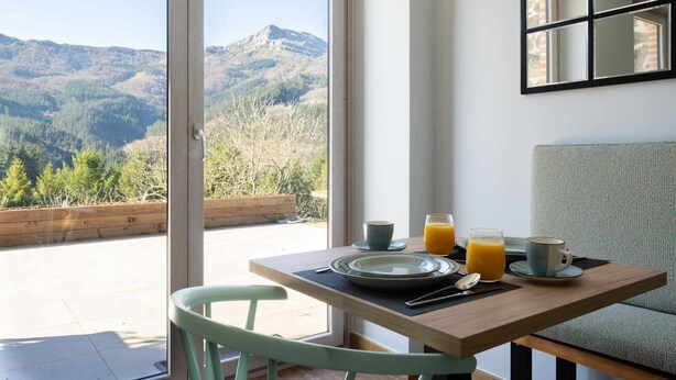 A dining area with a table set for two, a view of mountains, and a sofa. - Izadi (Zeanuri)