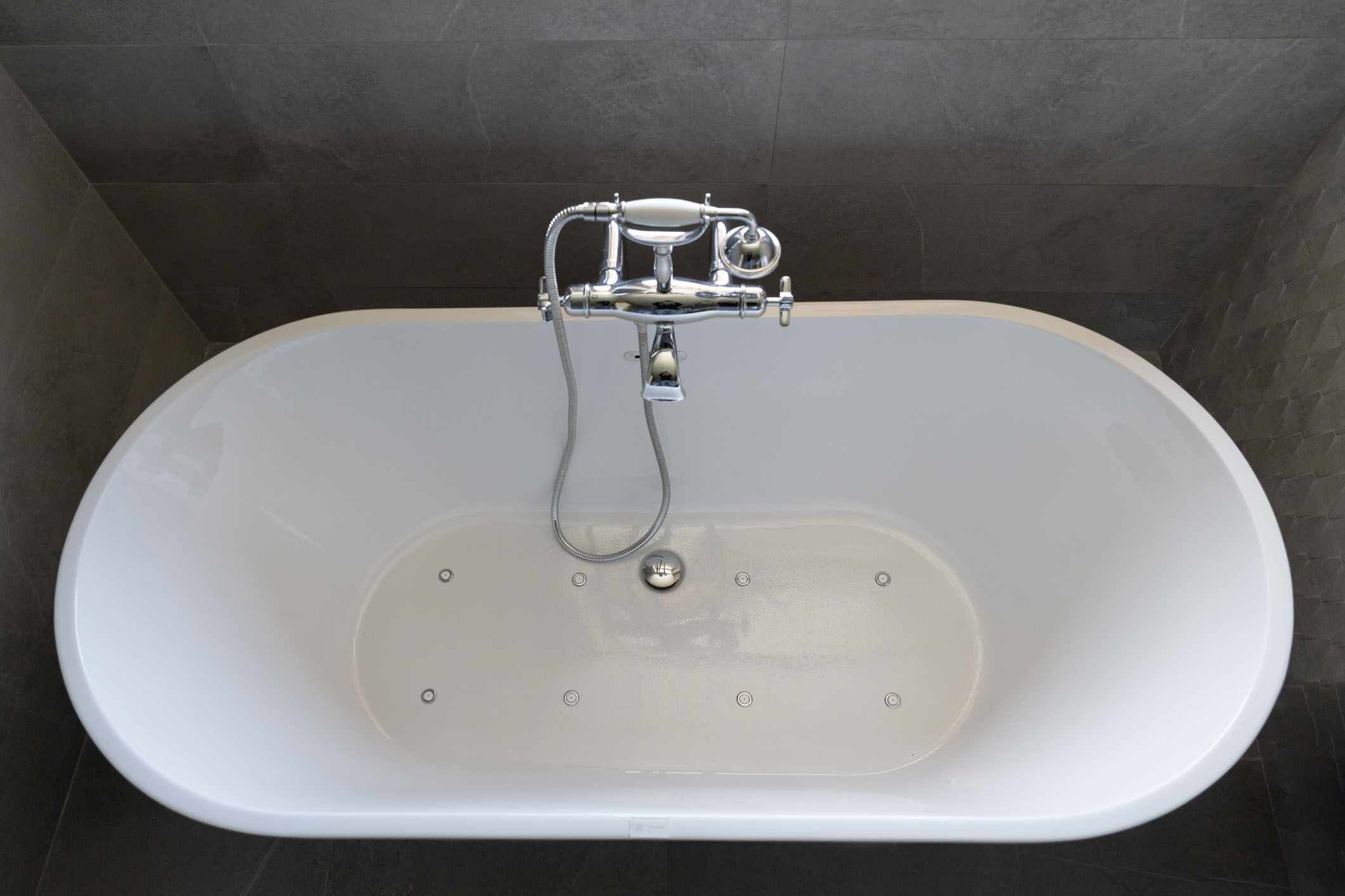 Eine moderne Badewanne mit chromfarbenem Wasserhahn und Duschkopf, vor einer dunkel gefliesten Wand.