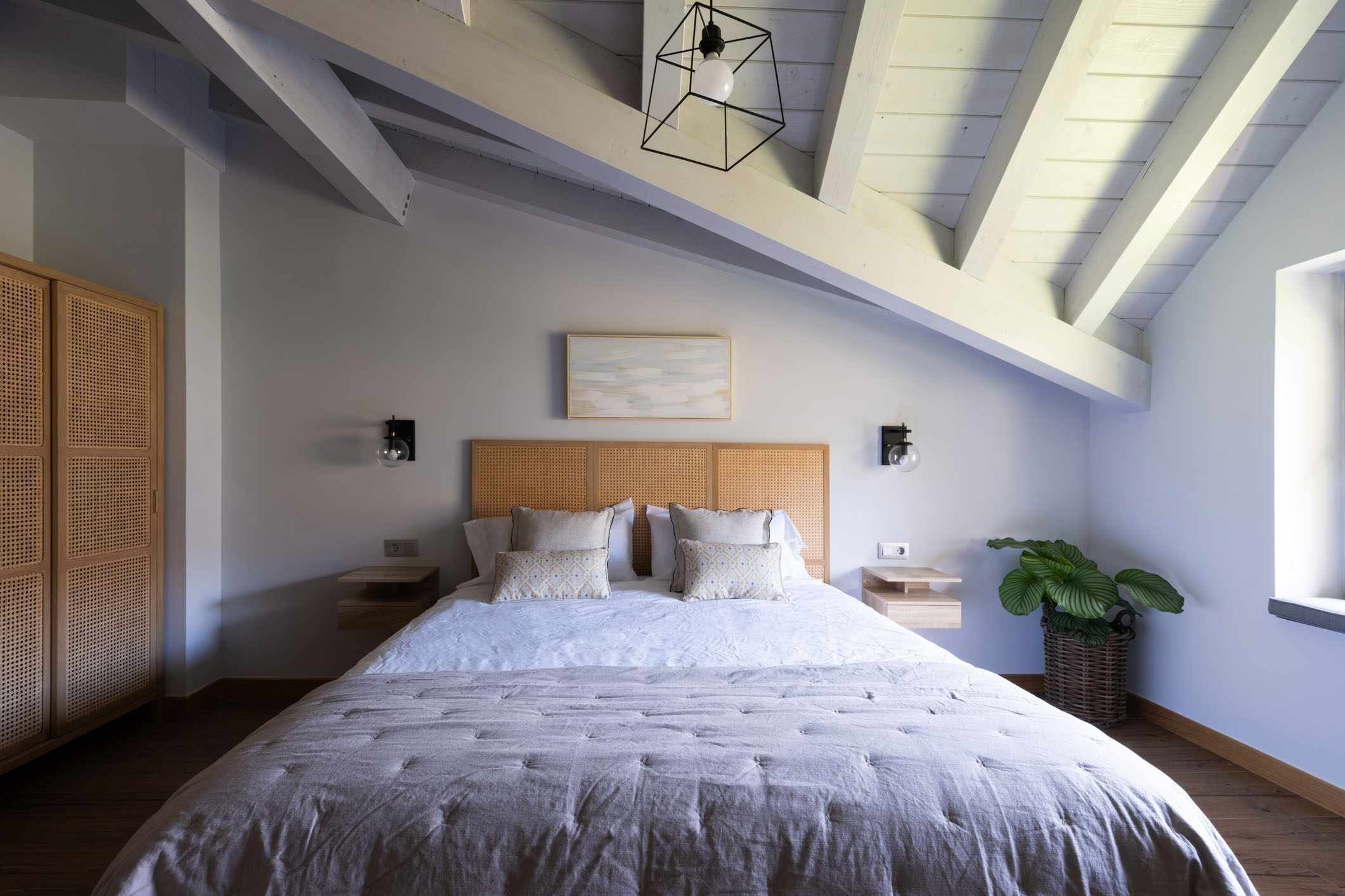 Un dormitorio con una cama grande, cabecera de madera, mesitas de noche, una lámpara de pared y una planta en maceta.