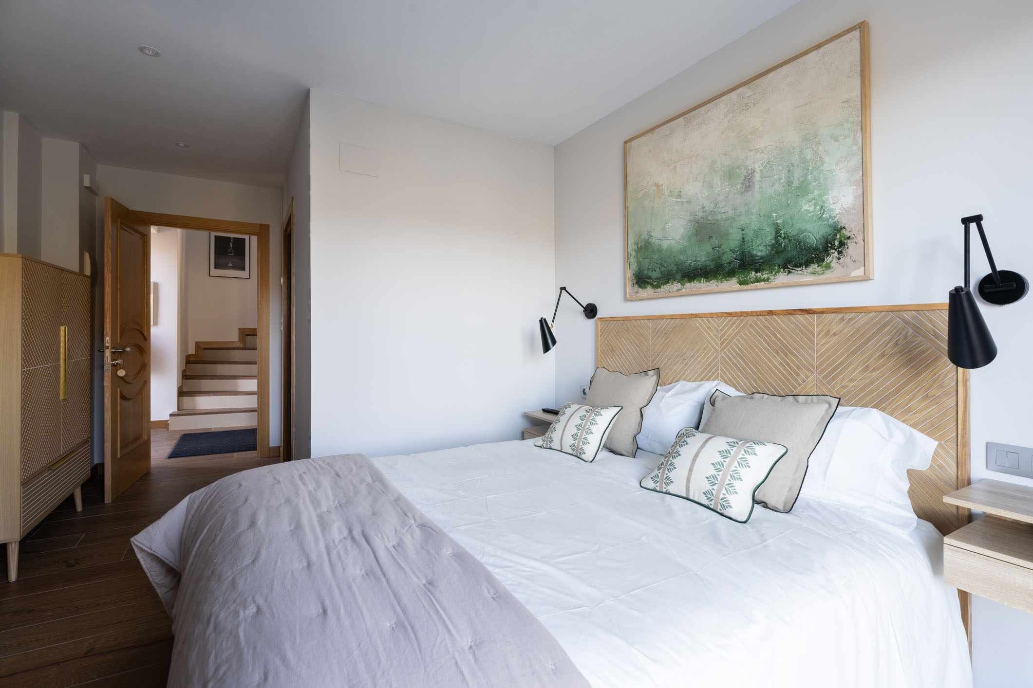 A bedroom with a large bed, a wooden headboard, a painting above the bed, and a nightstand with a lamp.