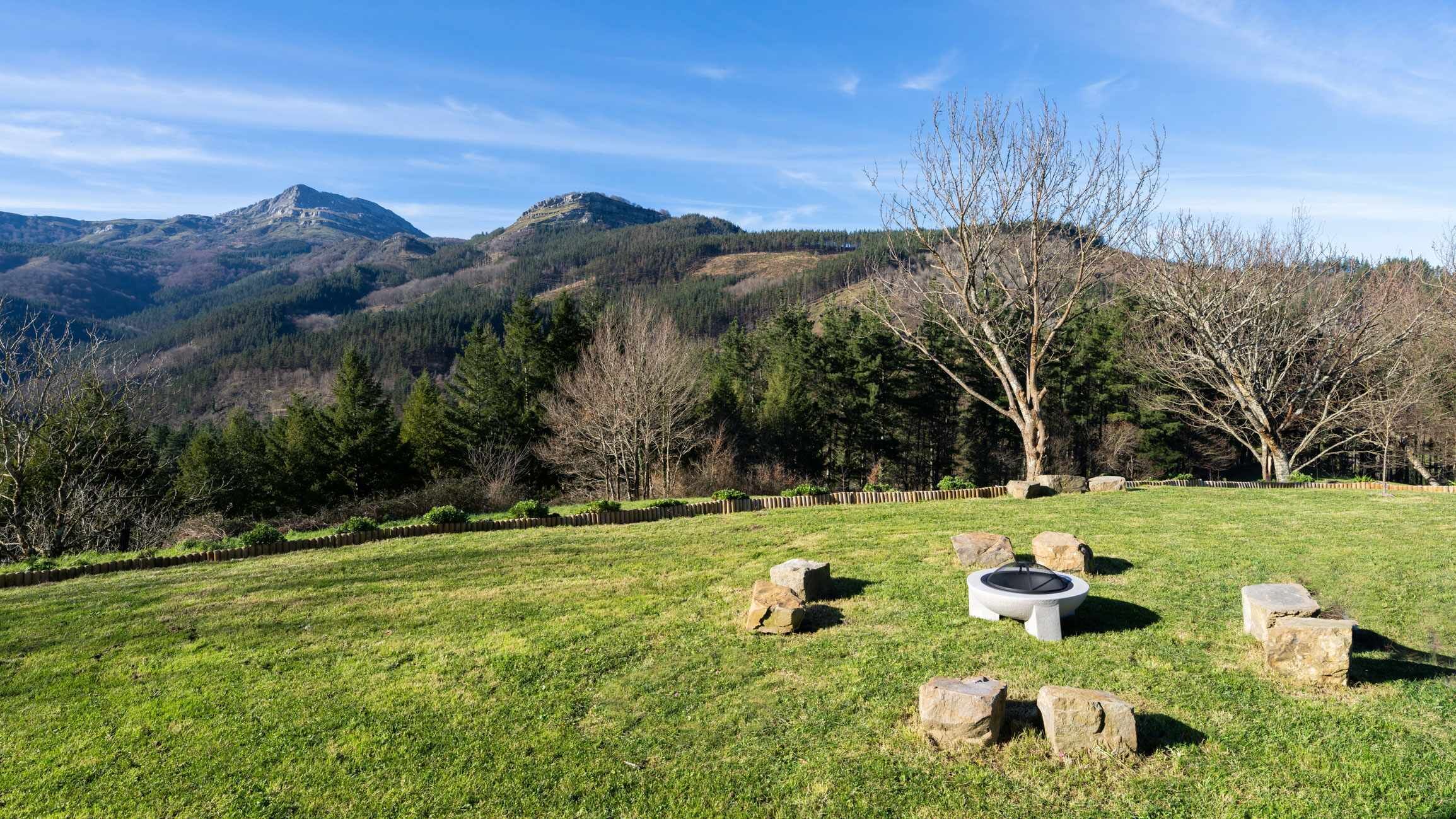 Un campo cubierto de césped con un hogar de piedra y zona de estar, rodeado de árboles y montañas al fondo.