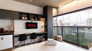 A modern hotel room with a large bed, a desk with two chairs, a TV displaying 'NETFLIX', and a city view through large windows.
