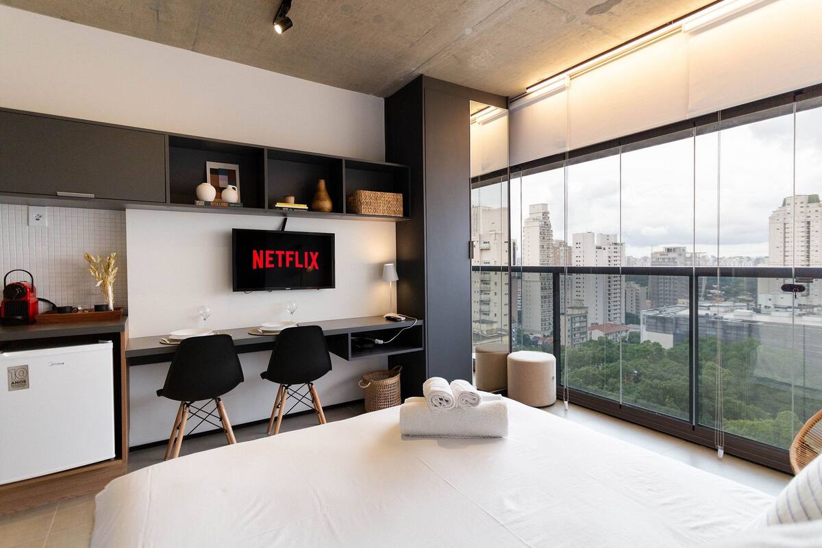 Quarto de hotel moderno com uma cama grande, uma escrivaninha com duas cadeiras, uma TV exibindo 'NETFLIX' e vista para a cidade através de amplas janelas.