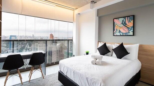 A modern hotel room with a large window offering a city view, a neatly made bed with white linens, and a small table with chairs.