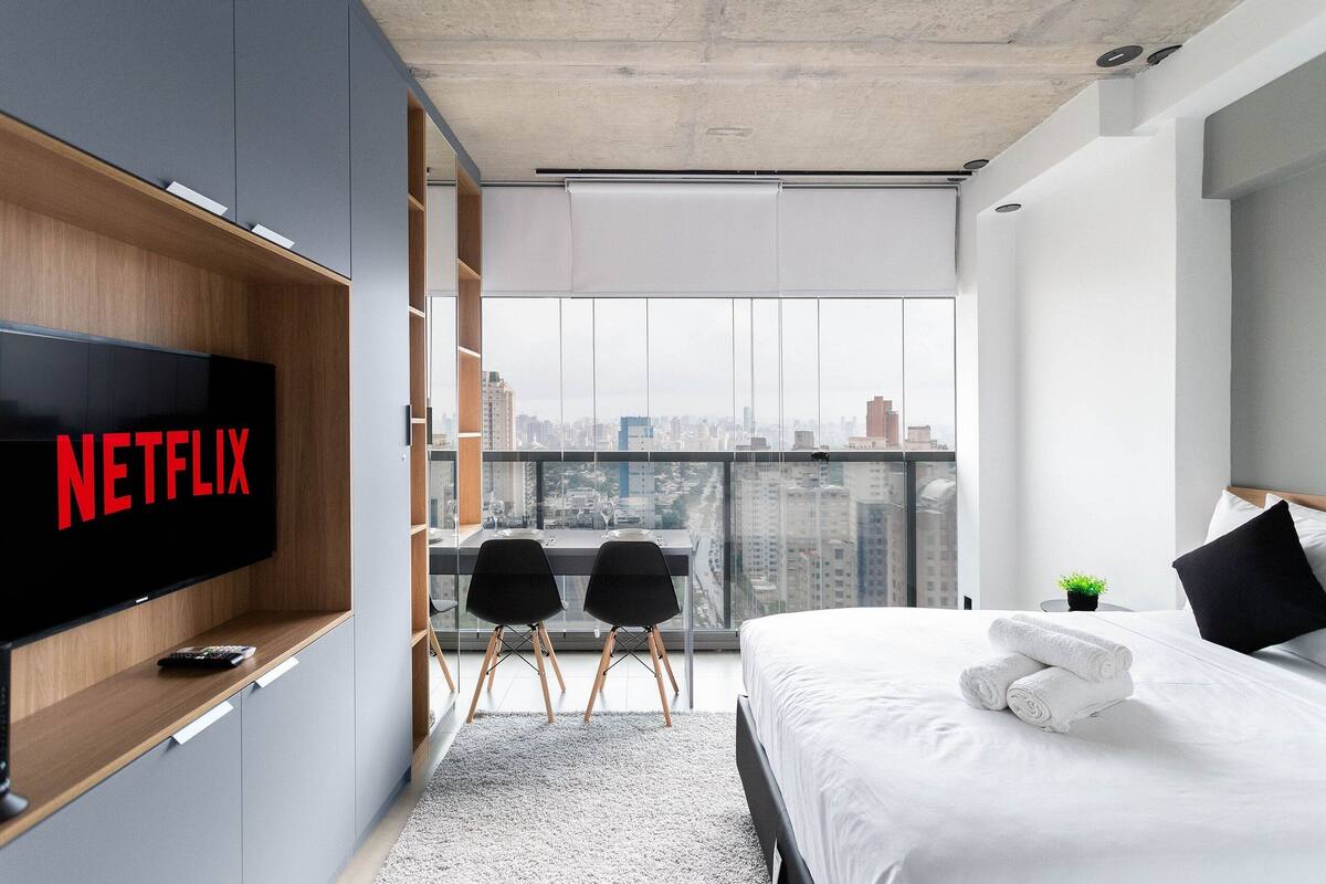 Quarto de hotel moderno com uma televisão grande exibindo 'NETFLIX', uma varanda com vista para a cidade, uma cama bem arrumada e uma pequena mesa com duas cadeiras.