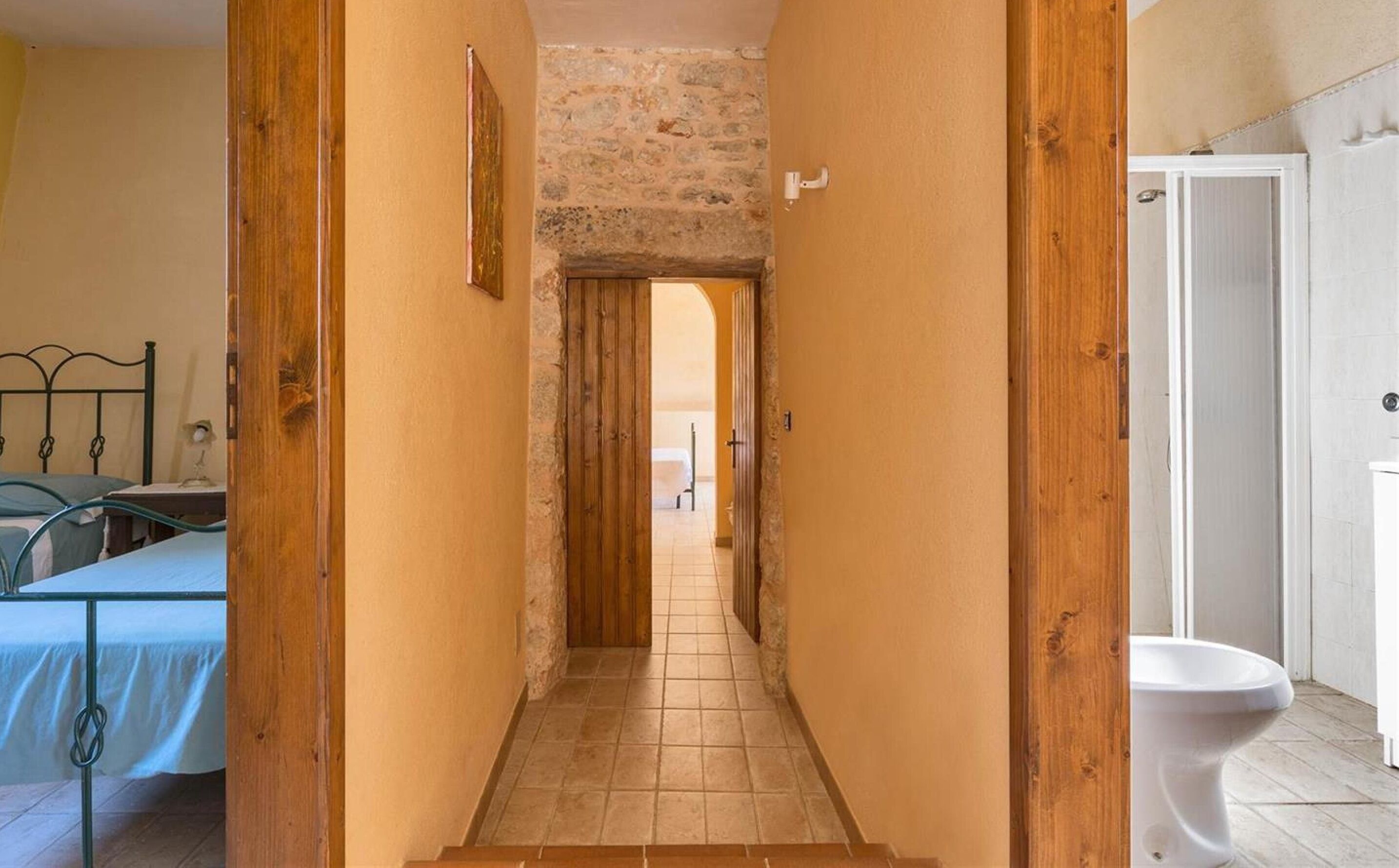 Un passadís estret amb portes de fusta, un llit amb estructura metàl·lica i un vàter blanc.