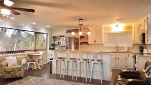 Une cuisine moderne avec des meubles blancs, un îlot central doté de tabourets, un coin repas avec une table et des chaises, et un ventilateur de plafond.