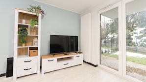 A modern living room with a flat-screen TV on a wooden TV stand, a white shelving unit with plants, and a sliding glass door leading to a garden. - Garden Facimiech 6a p4you pl (Kraków)