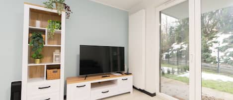 Una sala de estar moderna con un televisor de pantalla plana sobre un mueble de madera, una estantería blanca con plantas y un ventanal corredero que da a un jardín.