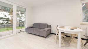 A modern living room with a grey sofa, a white dining table, and a large sliding glass door leading to a garden. - Garden Facimiech 6a p4you pl (Kraków)