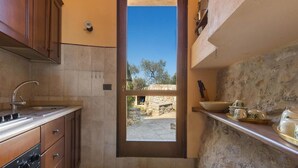 A compact kitchen with a stone wall, a wooden cabinet, and a window leading to an outdoor area. - 837 Masseria Colosso - Liama (Patu)