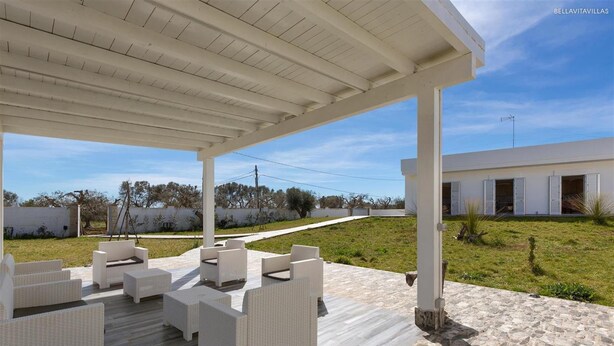 Une terrasse extérieure dotée de meubles en osier blanc et d’un sol en pierre, qui mène à un bâtiment moderne aux murs et volets blancs.