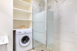 Un baño pequeño con lavadora, cabina de ducha de vidrio y paredes con azulejos de mármol.