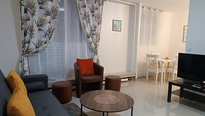 Una sala de estar con sofá, sillón, reposapiés y mesa de centro. Hay un televisor sobre un mueble bajo, una zona de comedor con mesa y sillas, y una ventana con cortinas ligeras.