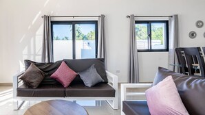 A modern living room with a dark brown sofa, a round wooden coffee table, and a white armchair. There are two windows with curtains, and a view of greenery outside. - Ultimate ’ Retreat with Pool & Beach Vibes!” (Sosúa)