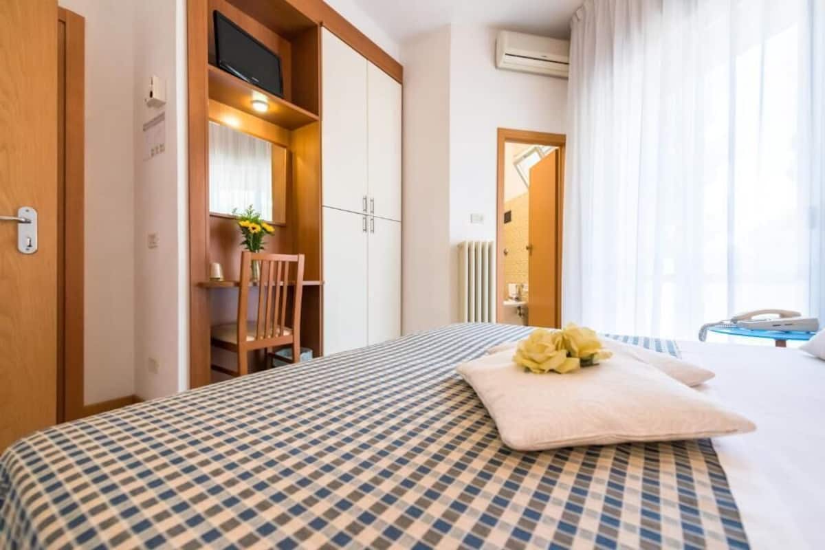 A hotel room with a checkered bedspread, a wooden chair, a television, and a window with curtains.