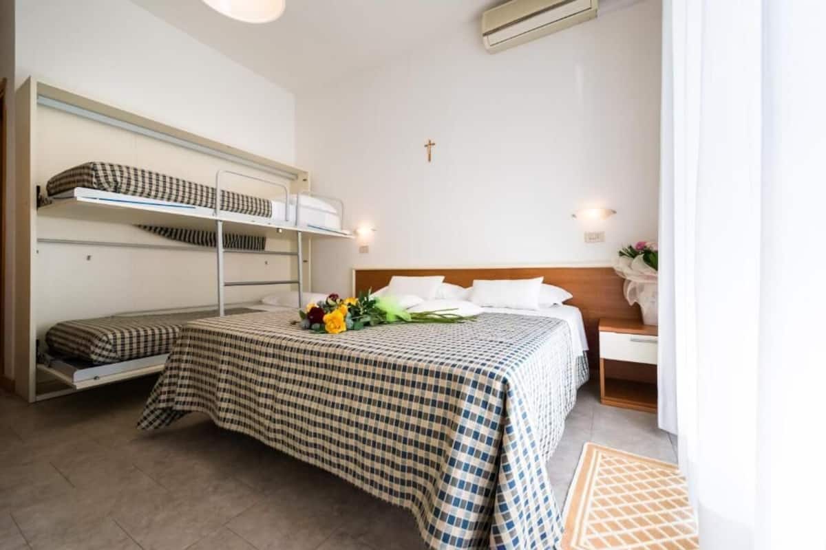 A room with a bunk bed, a single bed with a checkered bedspread, a bedside table with a lamp, and a wall-mounted air conditioner.