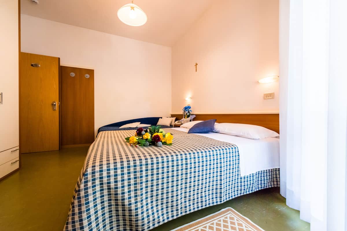 A hotel room with a checkered bedspread, a wooden door, a nightstand with a lamp, and a wall-mounted cross.