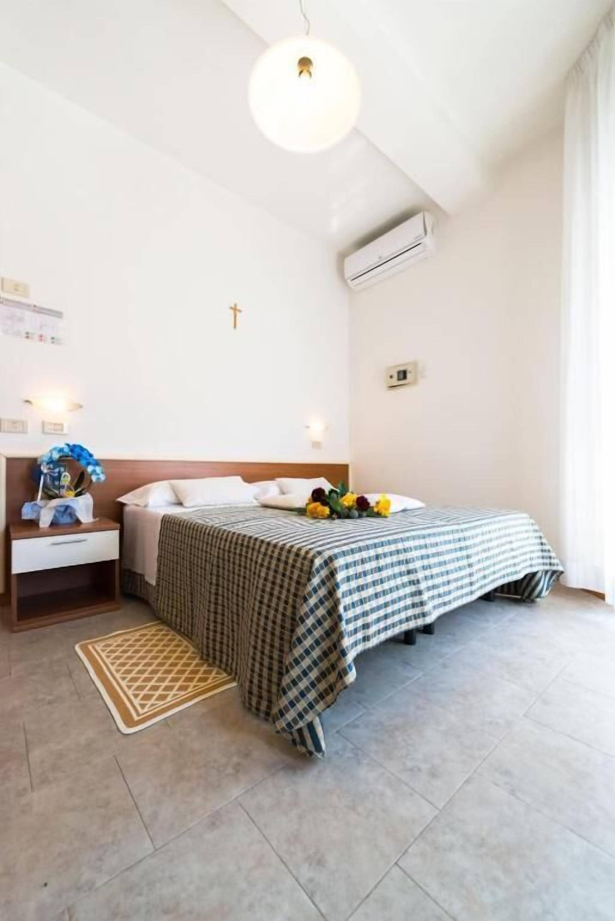 A hotel room with a bed, bedside table, lamp, air conditioning unit, and a wall-mounted cross.