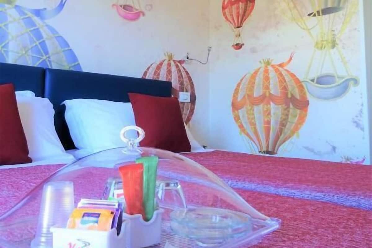 A hotel room with a bed covered by a clear plastic canopy, a bedside table with toiletries, and a wall decorated with hot air balloon murals.