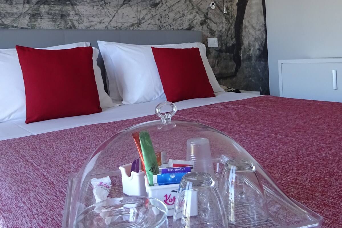 A hotel room with a bed, red and white pillows, a marble wall, and a glass-covered tray with toiletries.