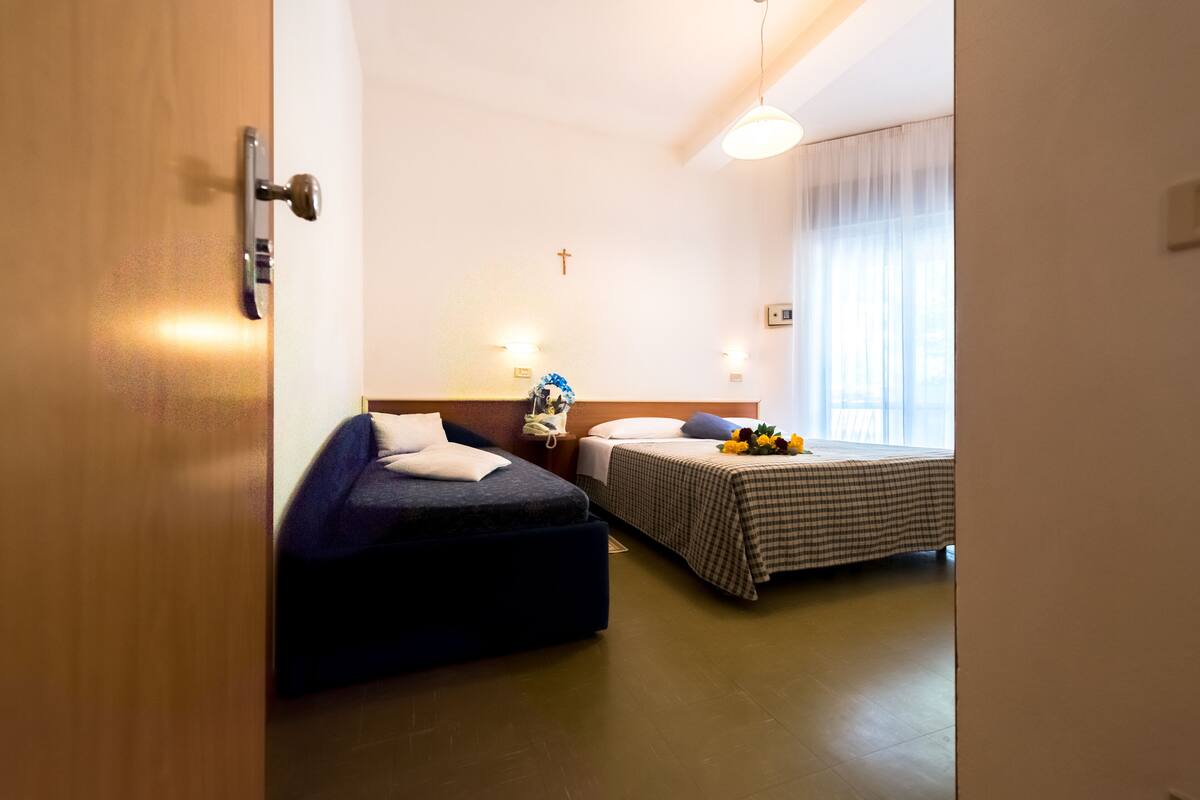 A hotel room with two beds, a wooden door, a cross on the wall, and a window with curtains.