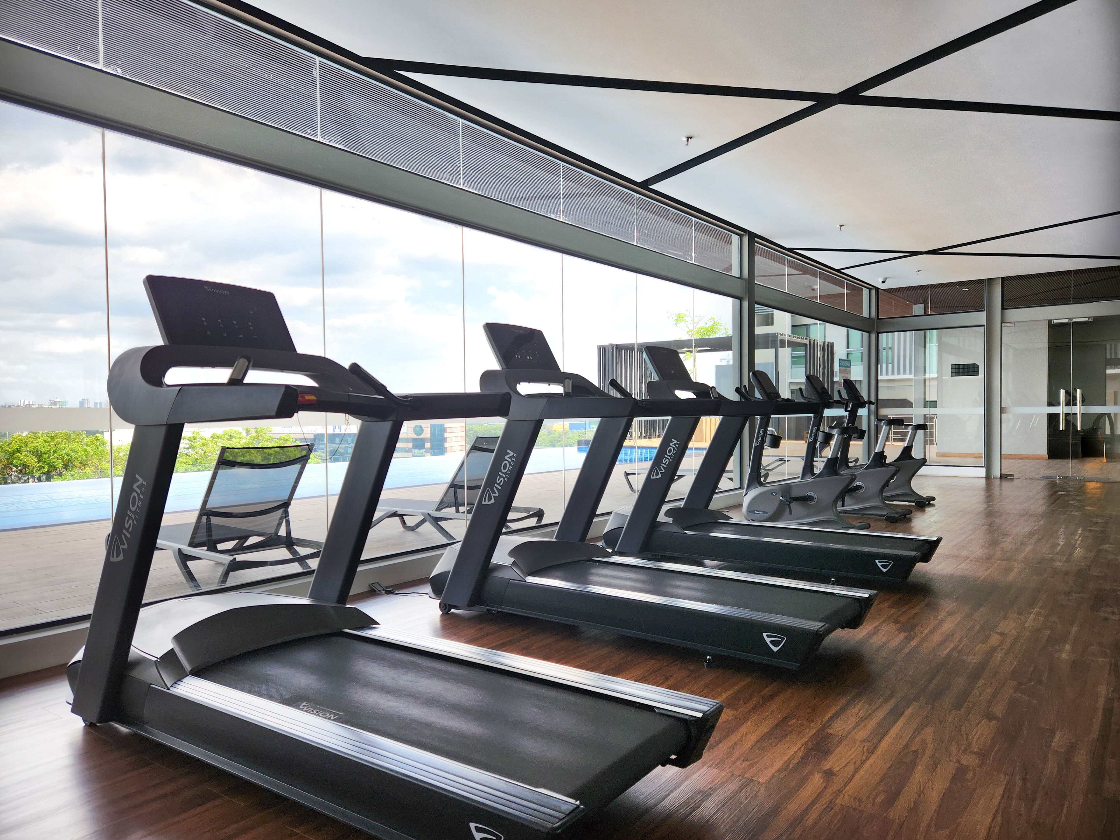 A gym with multiple treadmills aligned in a row, large windows, and a view of greenery and buildings.
