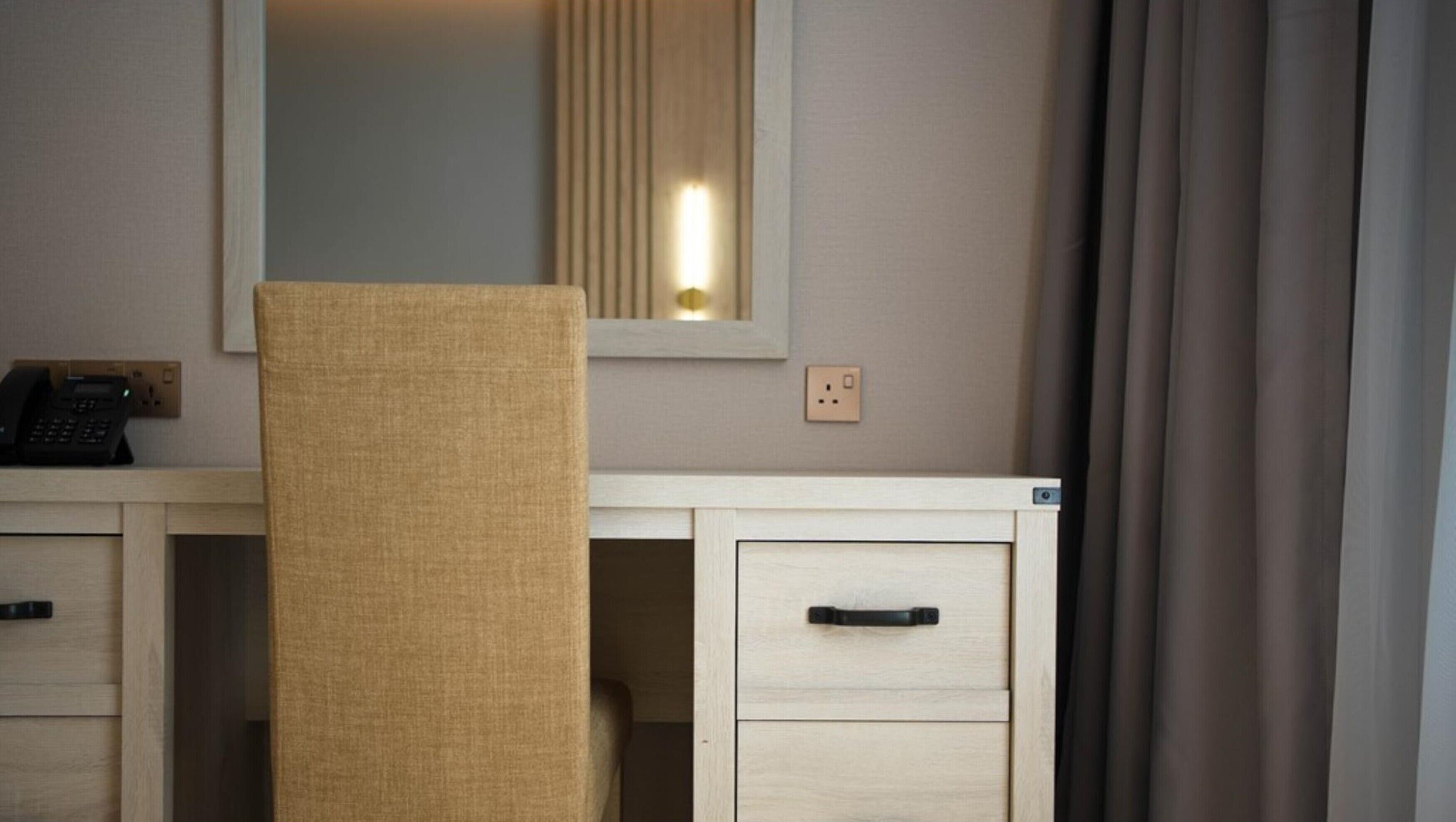 A hotel room with a desk, a chair, a telephone, a mirror, and a power outlet.