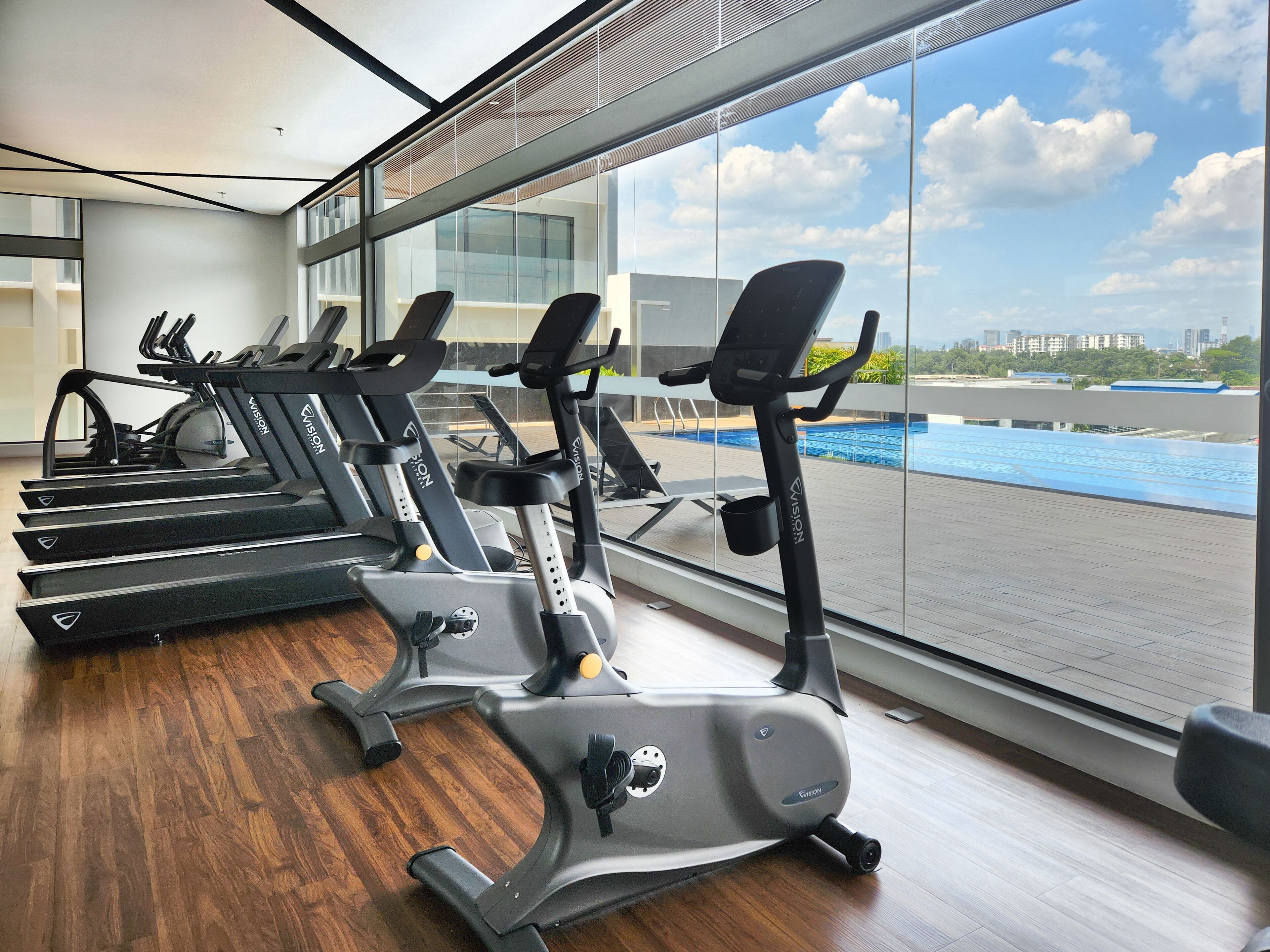 Et fitnesscenter med flere træningsmaskiner, store vinduer og udsigt til en swimmingpool og bygninger.